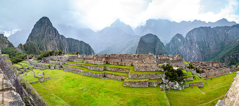 A complete scene of Macchu Pichu, in a blue sky day. Many tourists are present and scattered around the place visiting the ruins.