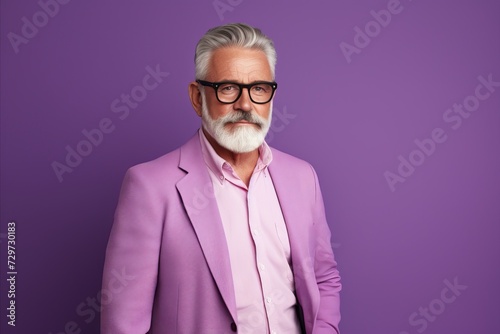 Portrait of a handsome senior man with gray beard and mustache wearing pink jacket and glasses, standing against purple background