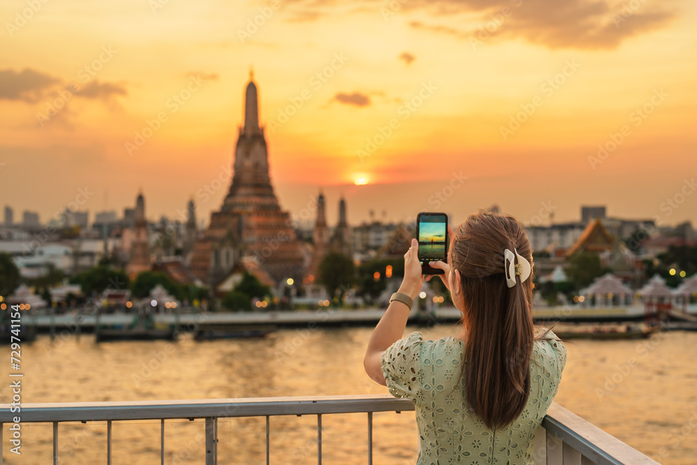 Obraz premium tourist woman enjoys view to Wat Arun Temple in sunset, Traveler take photo to Temple of Dawn by smartphone from rooftop bar. Landmark and Travel destination in Bangkok, Thailand and Southeast Asia