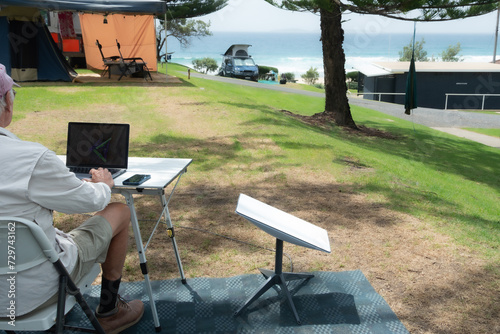 man with satellite internet dish connected working on computer at remote beach side campsite