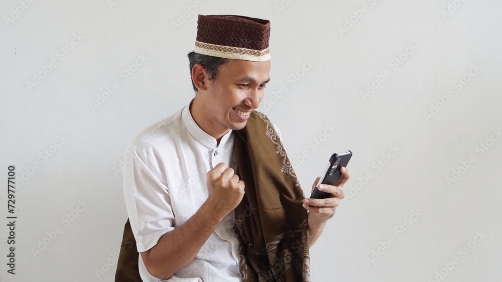 Fototapeta premium asian man with happy expression, playing mobile games, wearing cap and sarong