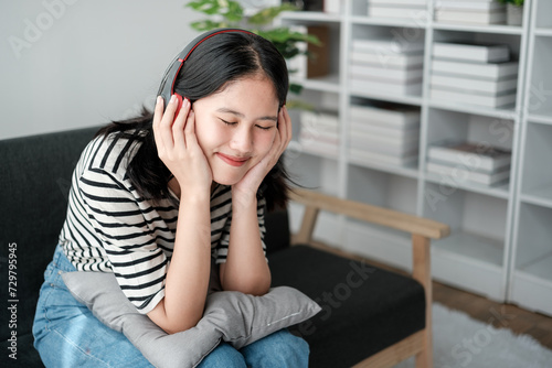 Attractive and happy young Asian woman in casual clothes wearing headphones, relaxes listening to music in her home office.