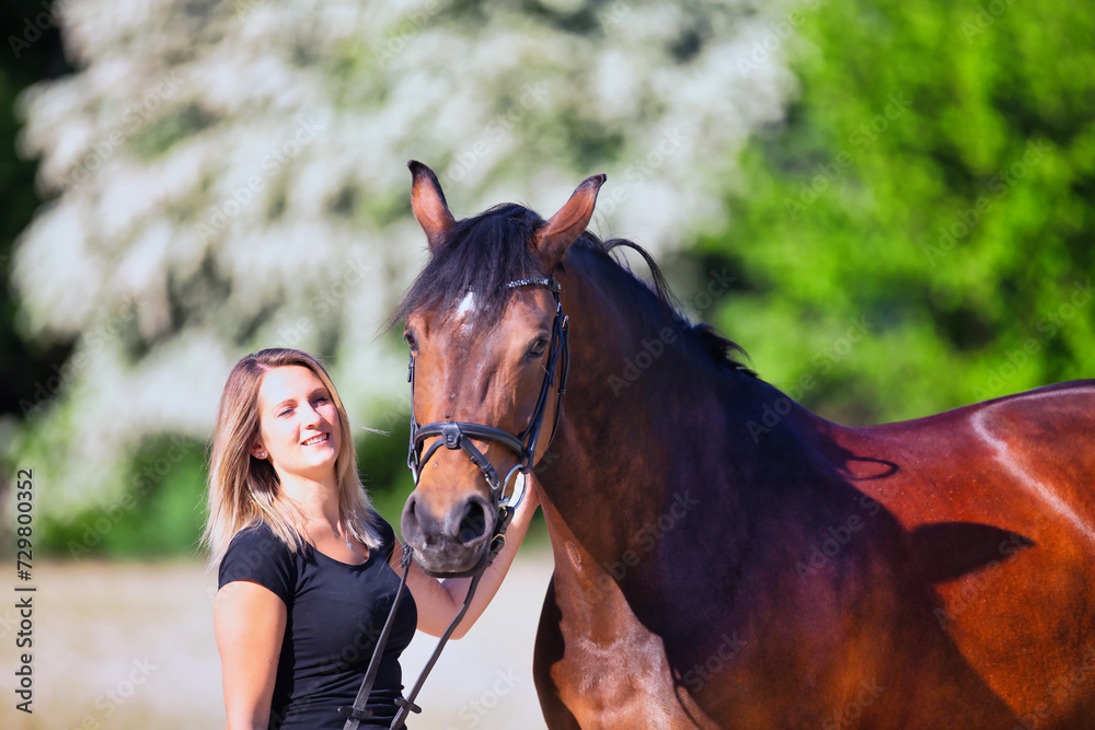Obraz premium Young brunette woman in the sunshine with her horse in front of white flowering bushes.