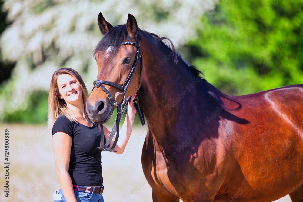 Obraz premium Young brunette woman in the sunshine with her horse in front of white flowering bushes.