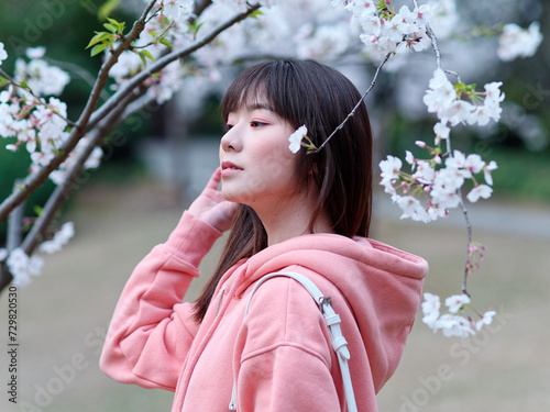 Beautiful young Chinese woman posing under blooming cherry tree, wearing pink loose oversized top with small white shoulder bag. Candid moment. Emotions, people, beauty, youth and lifestyle portrait.