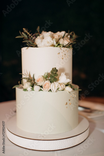 elegant white two-tiered wedding cake decorated with flowers on a small table against the backdrop of a wedding arch