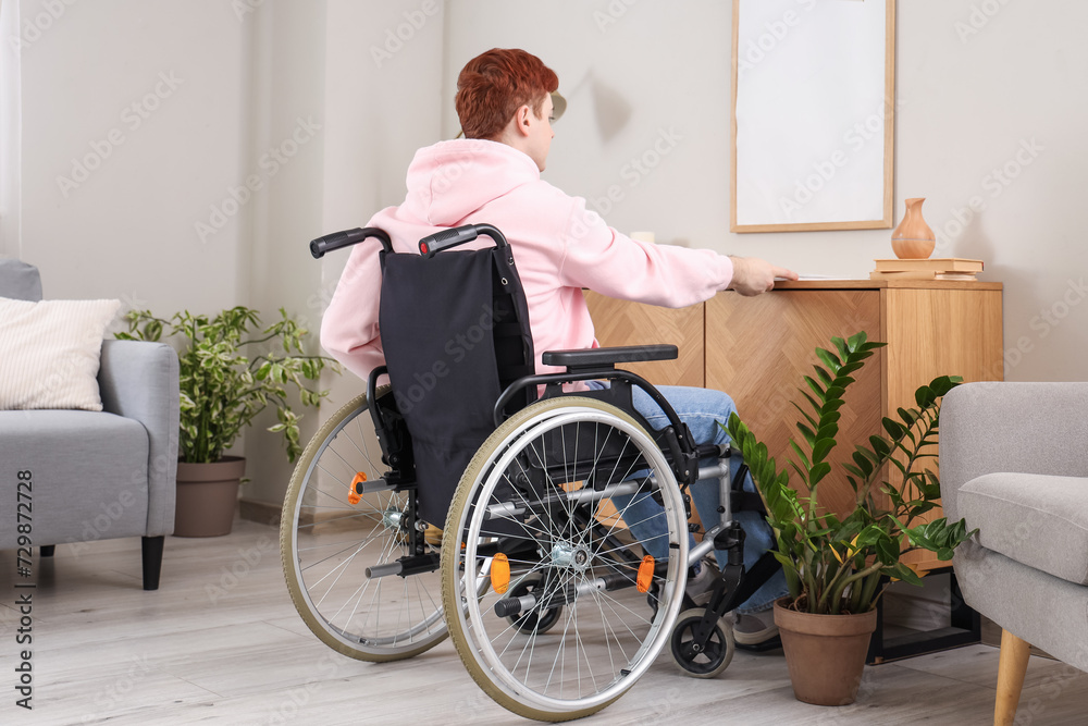 Young man in wheelchair taking magazine from shelf at home, back view Young man in wheelchair taking magazine from shelf at home, back view