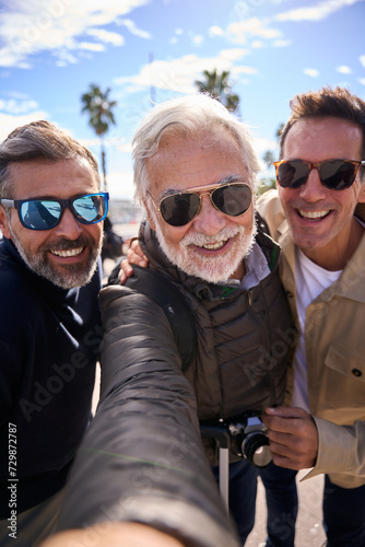 Vertical group diverse mature Caucasian handsome tourist men smiling happy take selfie with phone looking at front camera enjoying their vacation wearing sunglasses outdoors on a winter sunny day