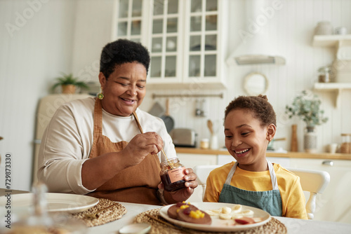 Cheerful African American grandma serving breakfast for her granddaughter adding jam to pancakes on plate photo