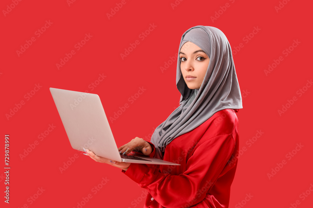Young Muslim woman in hijab using laptop on red background Young Muslim woman in hijab using laptop on red background