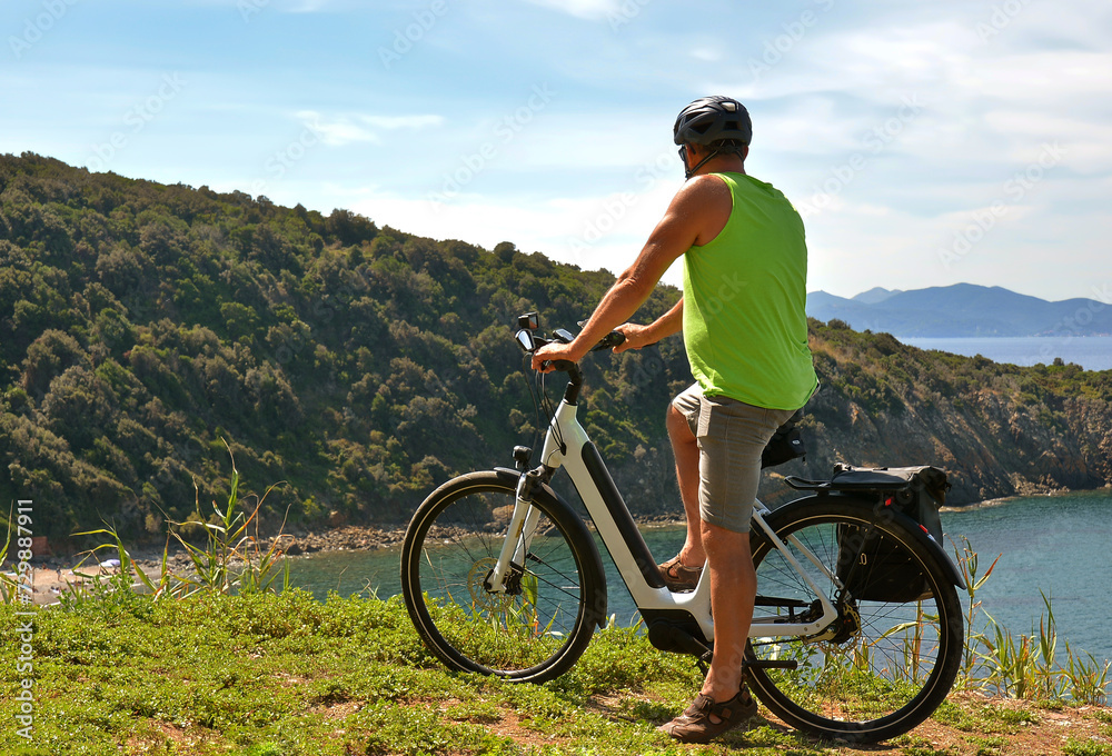 Obraz premium A middle-aged man in denim shorts and a green tank top on an electric bicycle stops and admires the beautiful scenery of the Italian coast of Tuscany during his cycling tour.