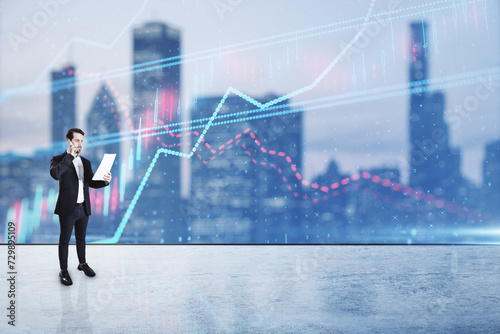 Young man holding document and talking on phone while standing on blurry city background with forex chart hologram. Success, finance, trade, future and tomorrow concept. Double exposure.