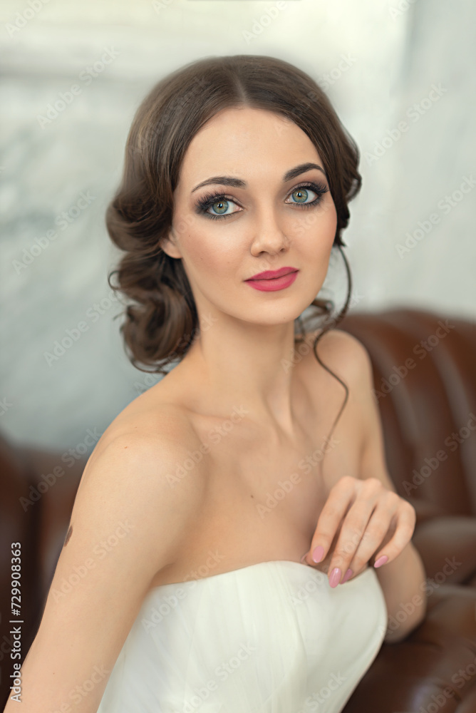 Portrait of a beautiful girl-bride in a wedding dress. A slender young woman with bare shoulders sits on the sofa. Wedding makeup and hairstyle. A beautiful face with big green eyes. Portrait of a beautiful girl-bride in a wedding dress. A slender young woman with bare shoulders sits on the sofa. Wedding makeup and hairstyle. A beautiful face with big green eyes.