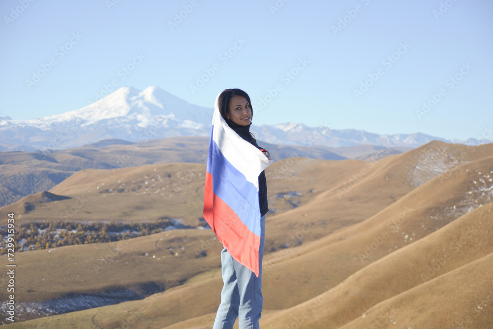 A young brunette woman stands against the backdrop of the snow-capped Mount Elbrus, looking at the camera, a Russian flag covers her shoulders. Tricolor against the backdrop of snow-capped