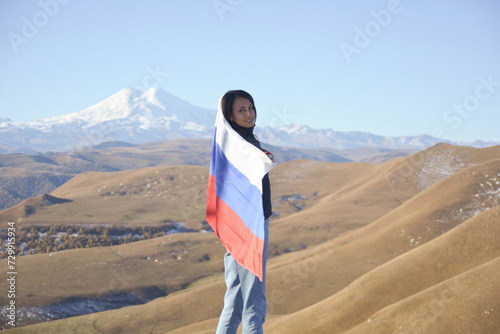 Obraz na płótnie A young brunette woman stands against the backdrop of the snow-capped Mount Elbrus, looking at the camera, a Russian flag covers her shoulders. Tricolor against the backdrop of snow-capped w salonie
