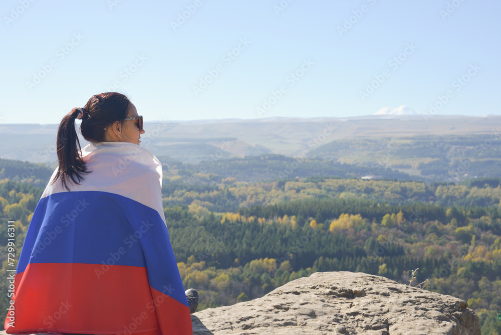A young brunette woman sits on a stone and looks at the mountains around, her shoulders are covered with a Russian flag.