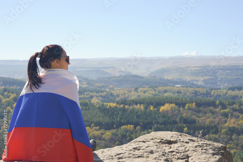 Obraz na płótnie A young brunette woman sits on a stone and looks at the mountains around, her shoulders are covered with a Russian flag. w salonie