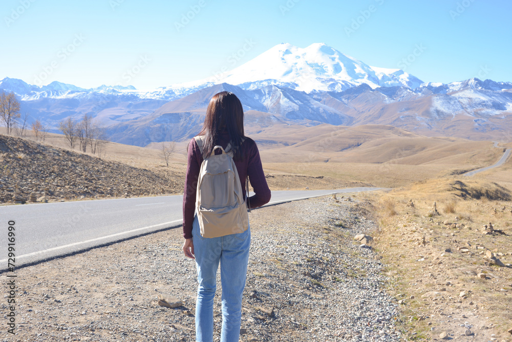 A female traveler with a backpack on her back admires the snow-covered Elbrus on a clear autumn day. View of Elbrus, North Caucasus, Russia
