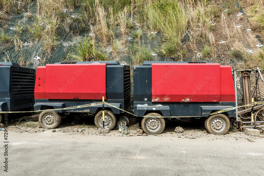 Row of large red industrial portable diesel engine generators stationed by the roadside with caution tape and metal fencing. Screw Air Compressor onsite at road and riprap construction.