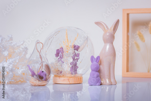 Easter bunnies standing next to a flower enclosed in a glass ball and a bird sitting in a glass house among boho-style decorations on a white tabletop and a light background