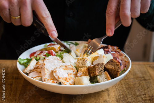 Close up of chicken Caesar salad with bacon, croutons, tomatoes, parmesan cheese, boiled eggs and dressing in a white bowl being eaten with knife and fork on a wooden table