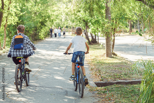 Two joyful boys riding bicycles on bicycle lane in park with lush foliage on warm and sunny summer day, enjoying freedom and beauty of outdoors