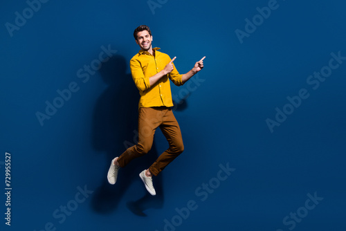 Full length photo of nice young male jumping point empty space dressed stylish yellow garment isolated on dark blue color background