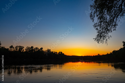 Great Don River in middle reaches. Orange sunset is reflected in water. Black silhouette of coastal forest. There are many trichopterans flying over smooth whirlpool current. Spring and high water © max5128