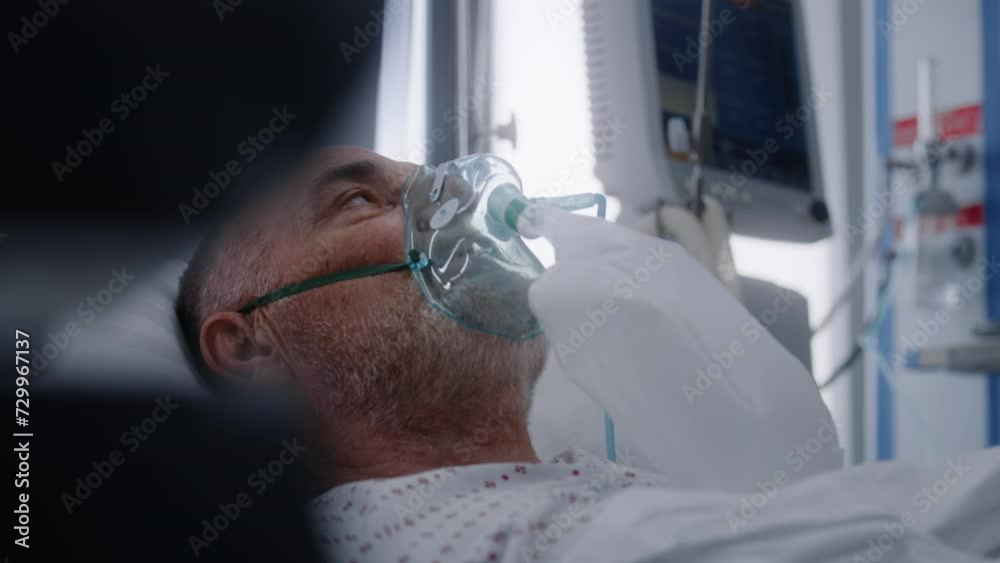 Old patient in oxygen mask lies in bed during lung ventilation. Nurse takes care of elderly man, uses monitor. Emergency room with modern equipment in hospital. Intensive care coronavirus department.