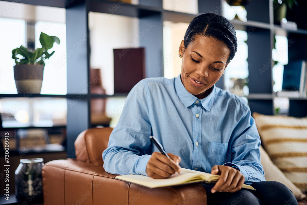 Obraz premium A smiling African woman writing some notes in her office, sitting on the sofa.