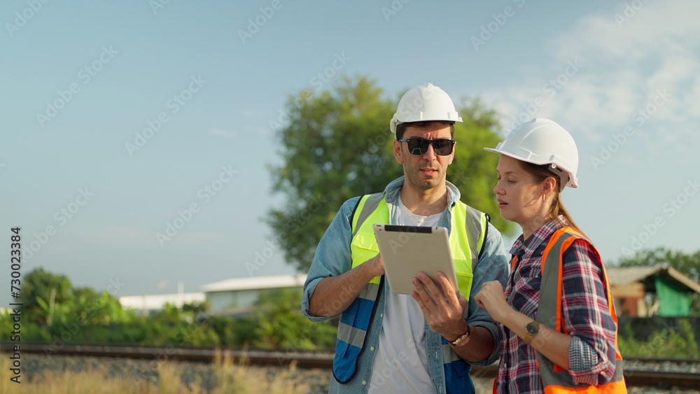 Two engineers in safety uniform and helmet using tablet discussion and inspection construction process railway while walking. Two engineer working on railway together. Teamwork concept Two engineers in safety uniform and helmet using tablet discussion and inspection construction process railway while walking. Two engineer working on railway together. Teamwork concept