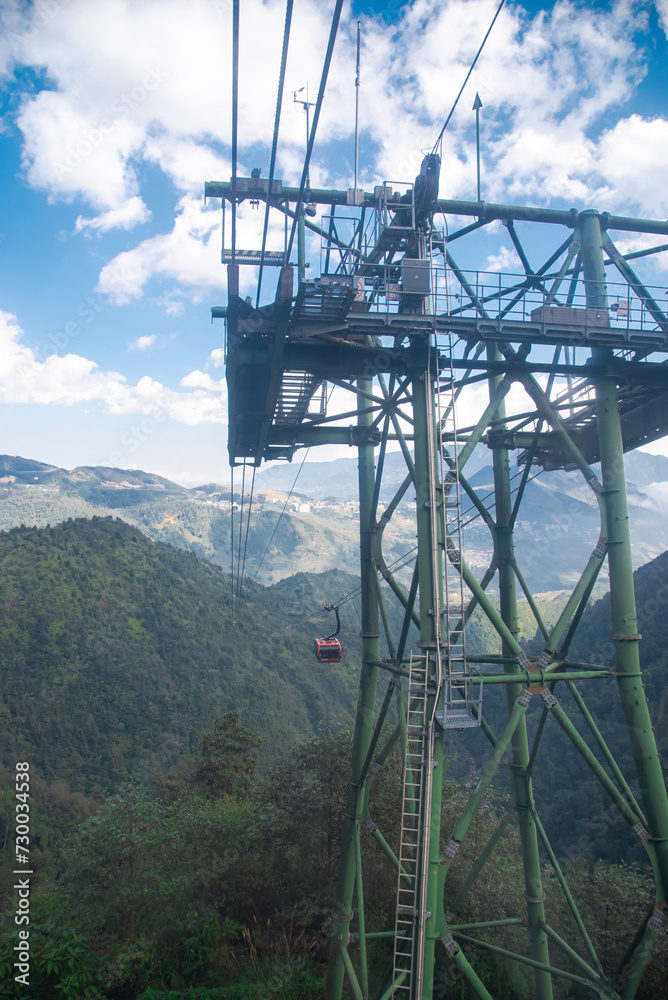 Close up steel framework of aerial lift pylon cable car pillar hauled gondola lift or ropeway above the ground, Hoang Lien Son mountain range lush green forest, sunny cloud blue sky in Sapa