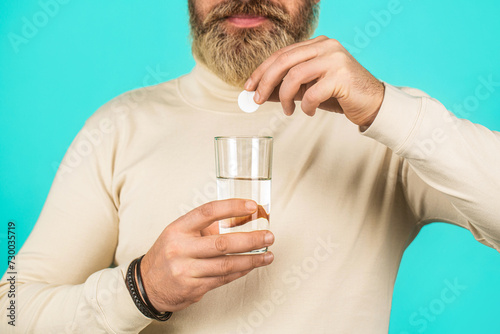 Man taking pill against headache. Male taking a pill with a glass of water. Man take some pills, holds glass of water, isolated on blue.Man taking drugs to releave headache photo