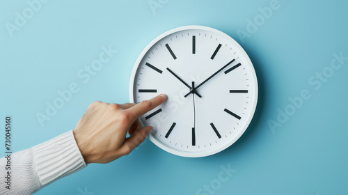 hand reaching out to a white wall clock on a light blue background, seemingly to adjust the time.