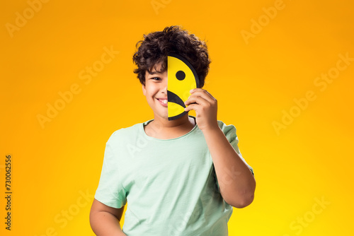 Child boy holds emoticons with sad emotions on half face.