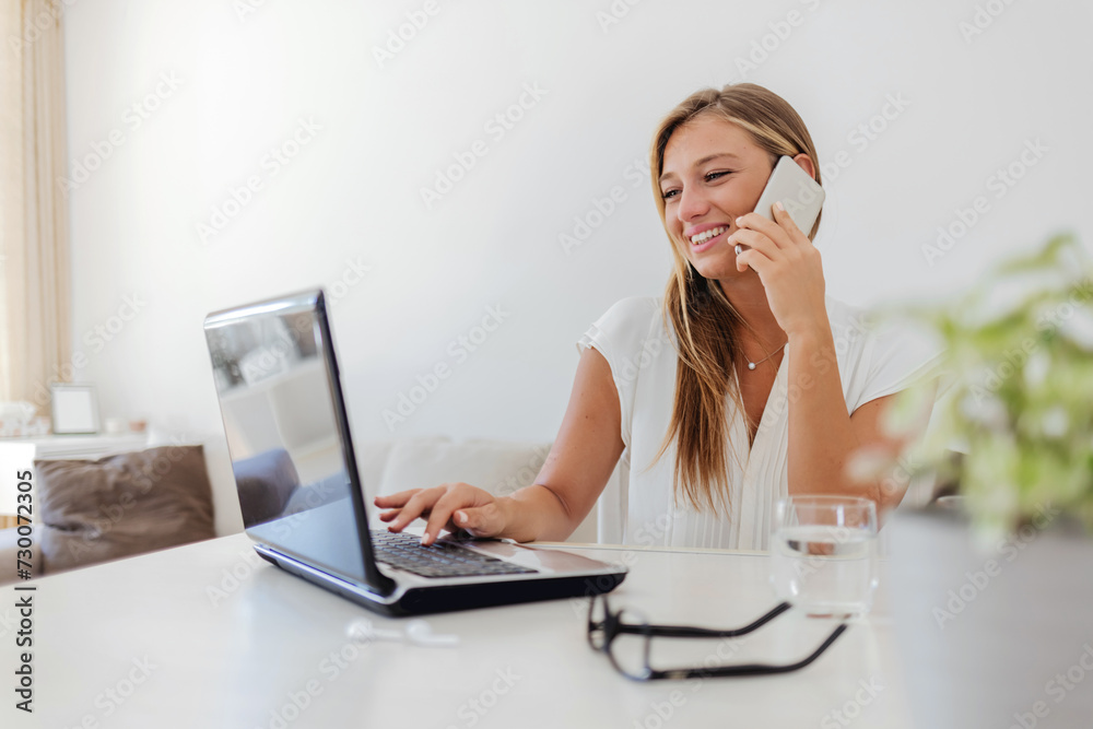 Attractive young businesswoman sitting in her home and talking on her cellphone while using her laptop. Happy businesswoman having a phone call while reading an e-mail on a computer at her home office Attractive young businesswoman sitting in her home and talking on her cellphone while using her laptop. Happy businesswoman having a phone call while reading an e-mail on a computer at her home office