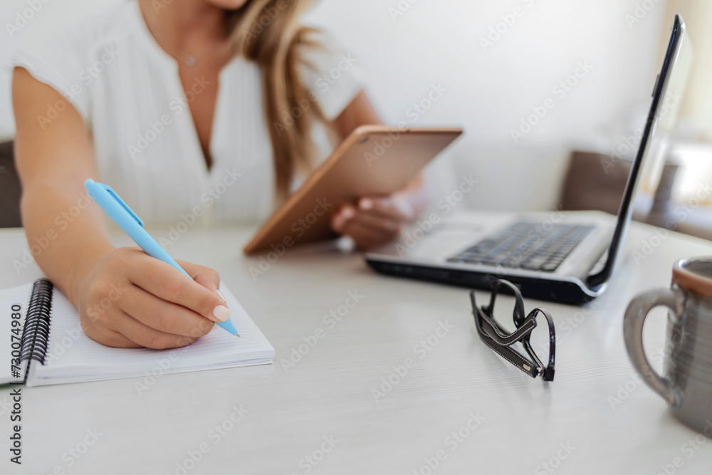 Φωτογραφία Beautiful smiling woman working from home, sitting at wooden desk with laptop and coffee, looking at digital tablet, writes something in a notebook