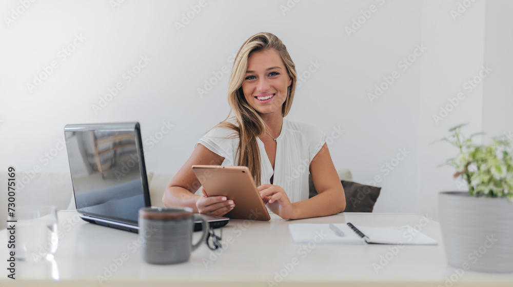 Portrait of smiling fulfilled young woman holding digital tablet device, looking at camera, sitting at desk at home with laptop computer in front of her and cup of coffee. Portrait of smiling fulfilled young woman holding digital tablet device, looking at camera, sitting at desk at home with laptop computer in front of her and cup of coffee.