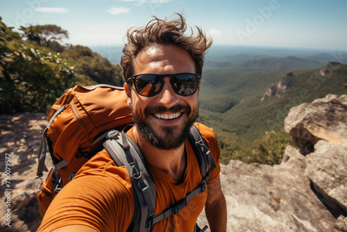 An athletic man in an orange t-shirt takes a selfie at the edge of a mountain, with a panoramic view of green mountains and valleys forming the backdrop