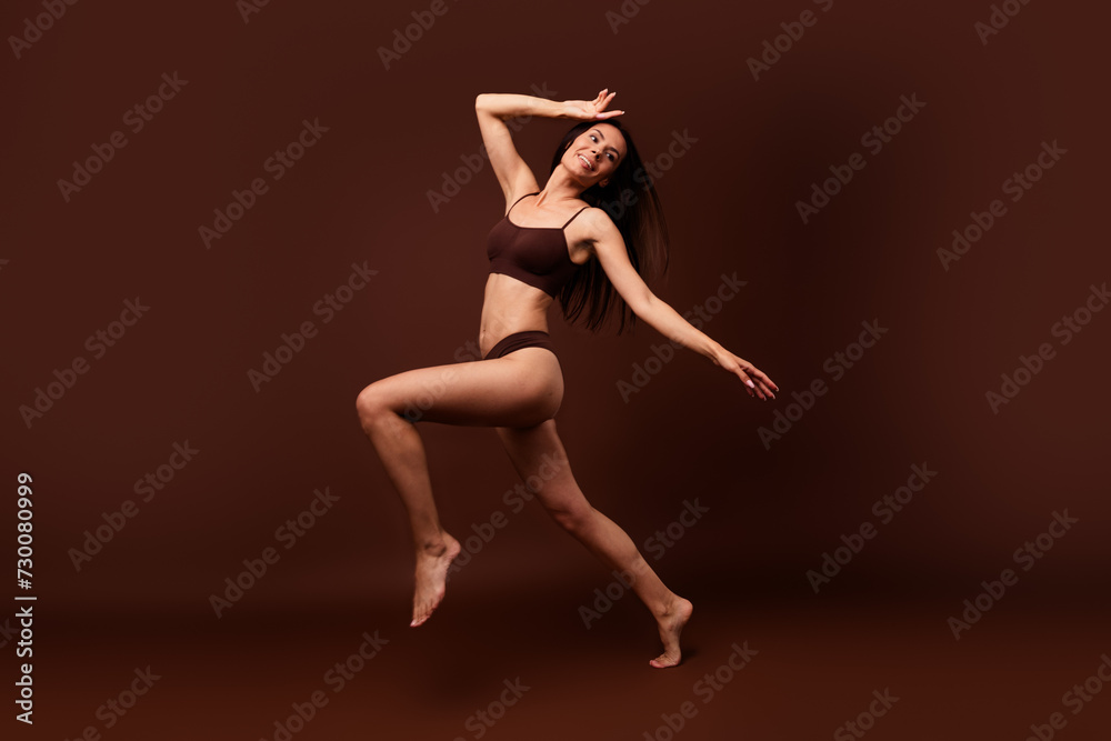 Unfiltered full length photo of sensual girl stand on one leg hands on head look empty space isolated on dark brown color background