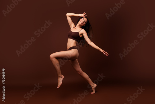 Unfiltered full length photo of sensual girl stand on one leg hands on head look empty space isolated on dark brown color background © deagreez
