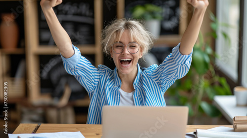 exuberant woman with tousled hair and glasses is raising her arms triumphantly in front of a laptop, expressing joy and success