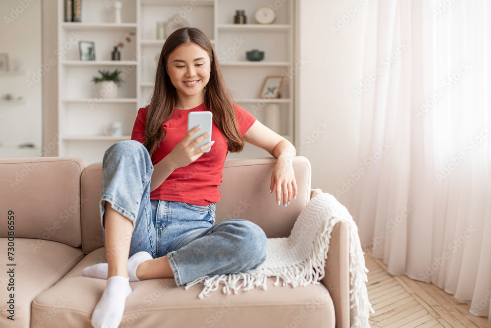 Relaxed young asian woman sitting on couch and looking at her smartphone Relaxed young asian woman sitting on couch and looking at her smartphone