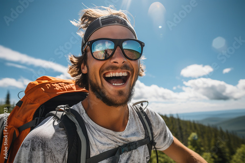 Shot from low angle of a man in a light grey t-shirt stands amidst lush green trees under clear skies, with an orange backpack suggesting a travelling adventure © Óscar