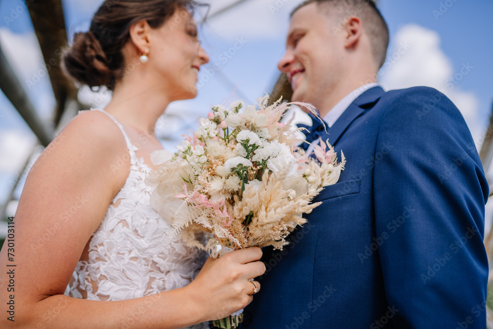 Valmiera, Latvia - July 7, 2023 - Close-up of a bride and groom on their wedding day, holding a bouquet, under a blue sky.