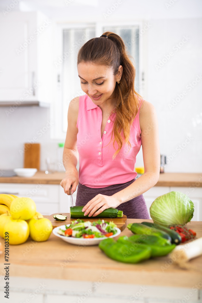 Positive young girl slicing vegetables for salad in cozy kitchen Positive young girl slicing vegetables for salad in cozy kitchen