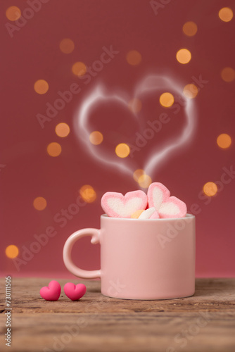 hot drink with heart shape smoke on pink coffee cup with heart shape marshmallow on top, with two small heart placed side by side on wooden table, bokeh light at background. romantic love relationship