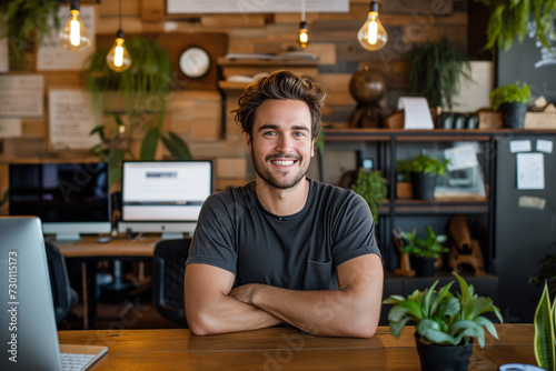 Happy Caucasian young male freelancer smiling while seated in a co working space with greenery, Wooden office interior, handsome young man seated in a modern interior background with plants around