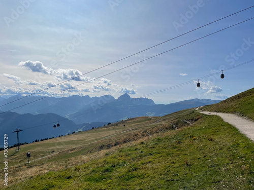 summer panoromic view of a hiking path through green mountain meadows with a gondola cableway and a hazy mountain range in the distance