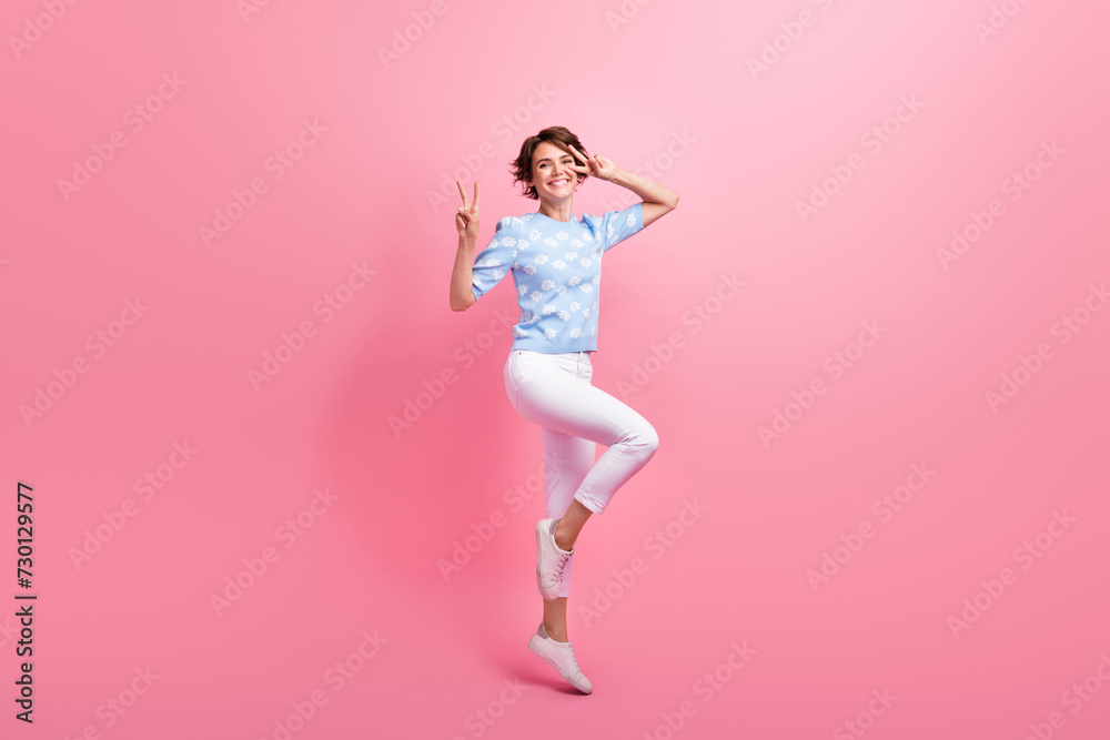 Full size portrait of pretty active girl jump demonstrate v-sign near eye have good mood isolated on pink color background